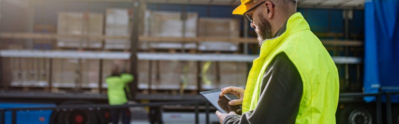 Warehouse manager overseeing unloading of truck, holding tablet, looking at cargo details, checking delivered items, goods against order, quality control.