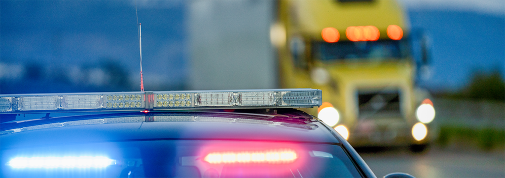 Police car with flashing red and blue lights on a road, a yellow truck approaching in the blurred background.